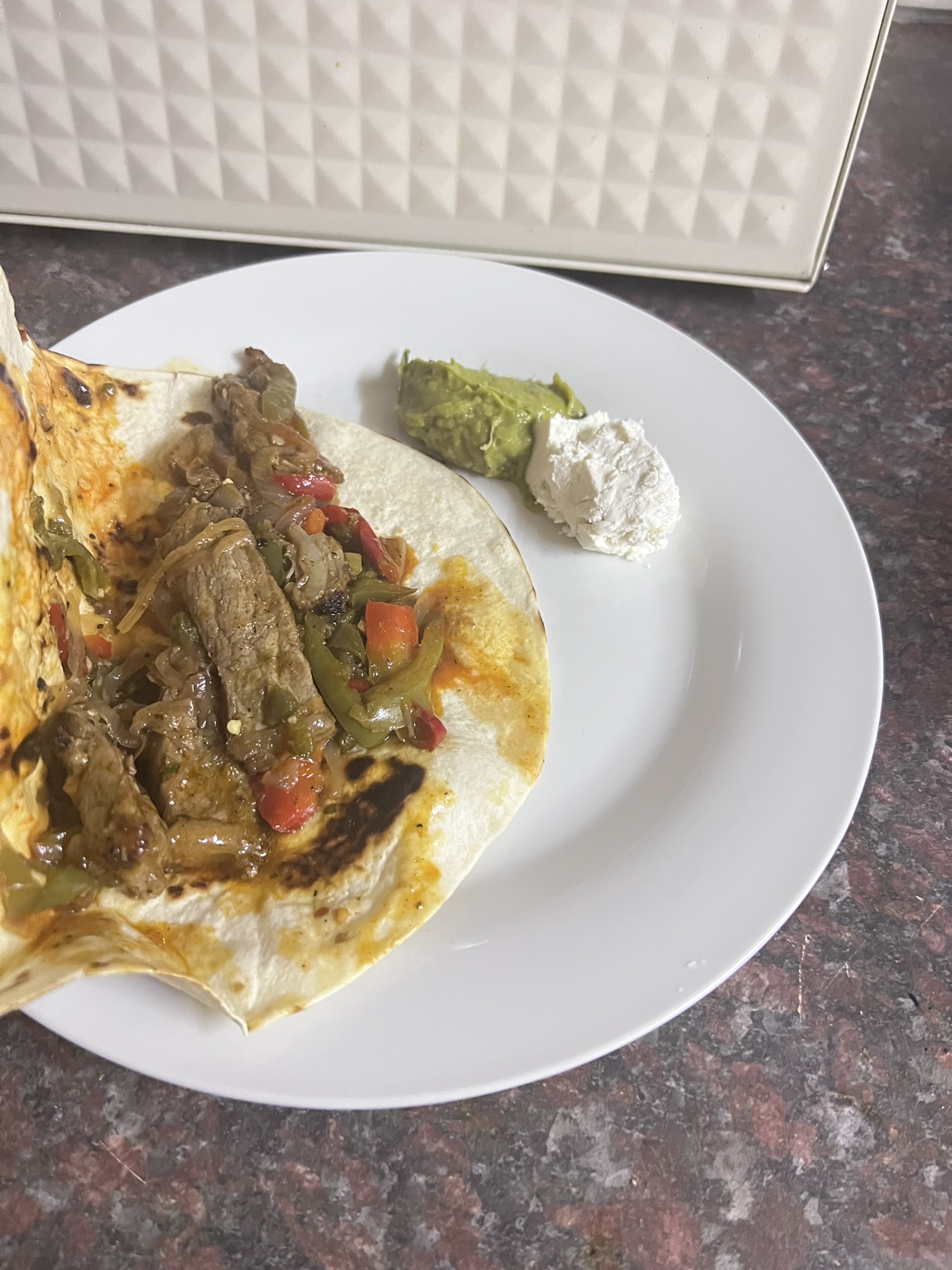 Open tortilla loaded with steak strips, charred red and green peppers and onion, with guacamole and sour cream alongside